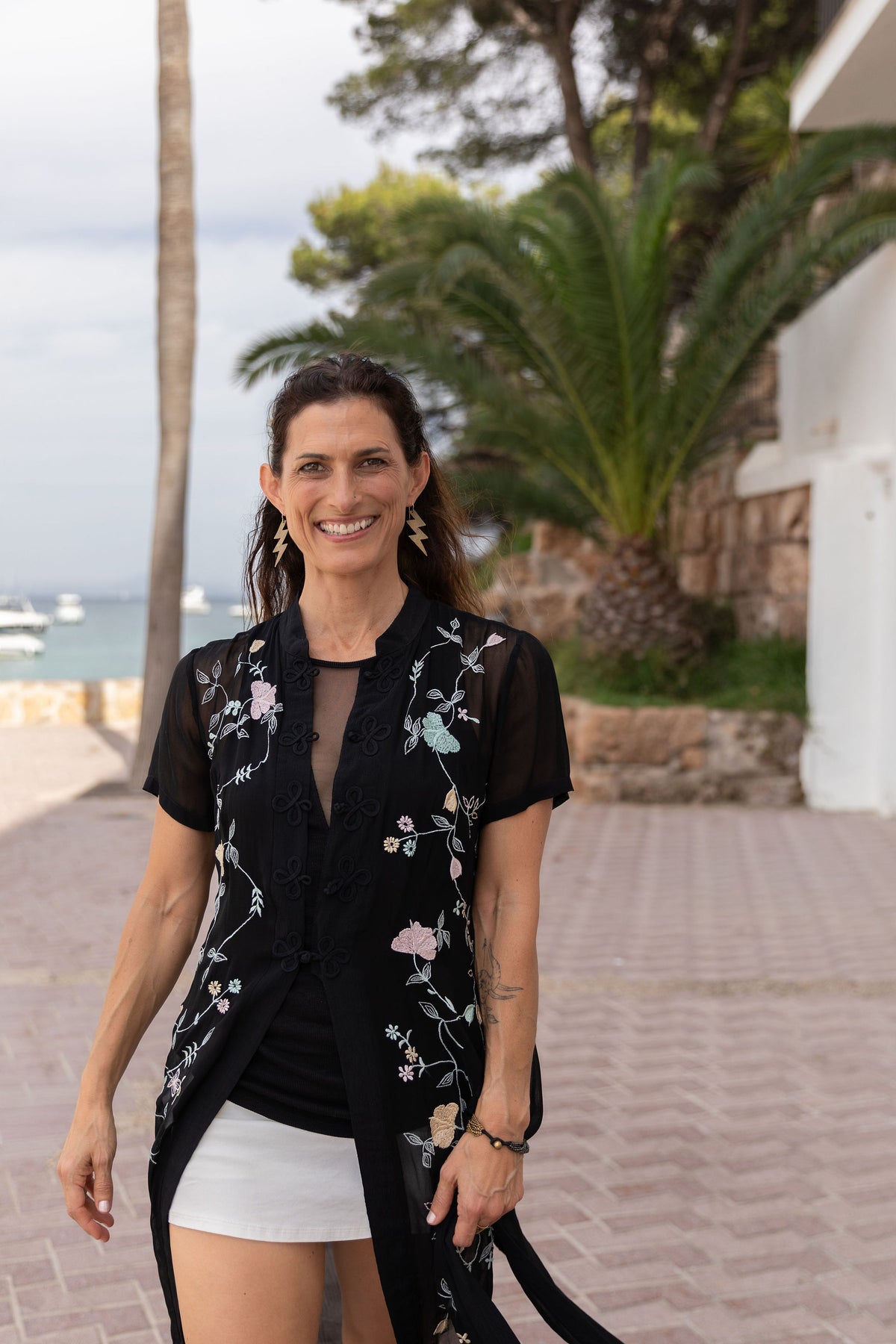 Dr. Katie Milano smiles at the camera at the seaside while wearing a sheer black short-sleeve cardigan with light pink and blue floral embroidery over a black top and white skirt. 