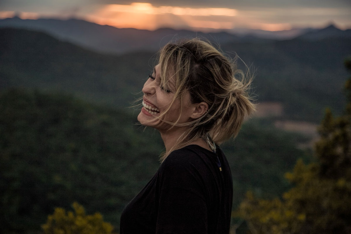 A woman with blonde hair tied back is shown in profile, smiling and looking upward against a mountain landscape at sunset. She is wearing a black top.